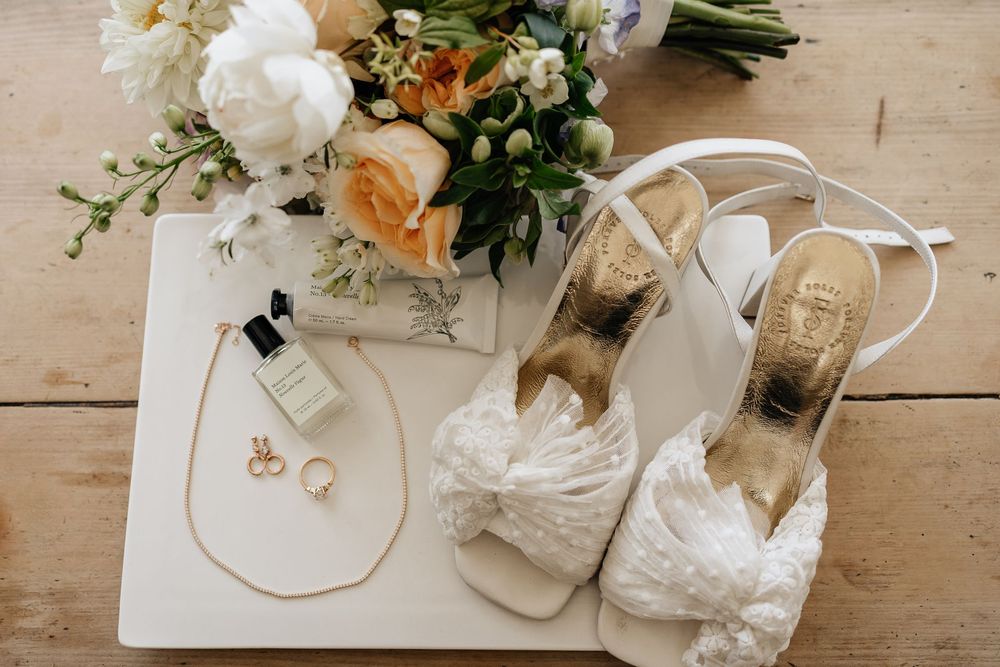 Wedding accessories including white bridal shoes, perfume, jewelry and floral bouquet arranged on wooden surface.