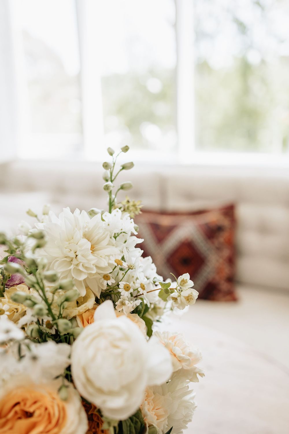 Elegant floral arrangement with white dahlias and peach roses beside a decorative pillow in a bright window setting.