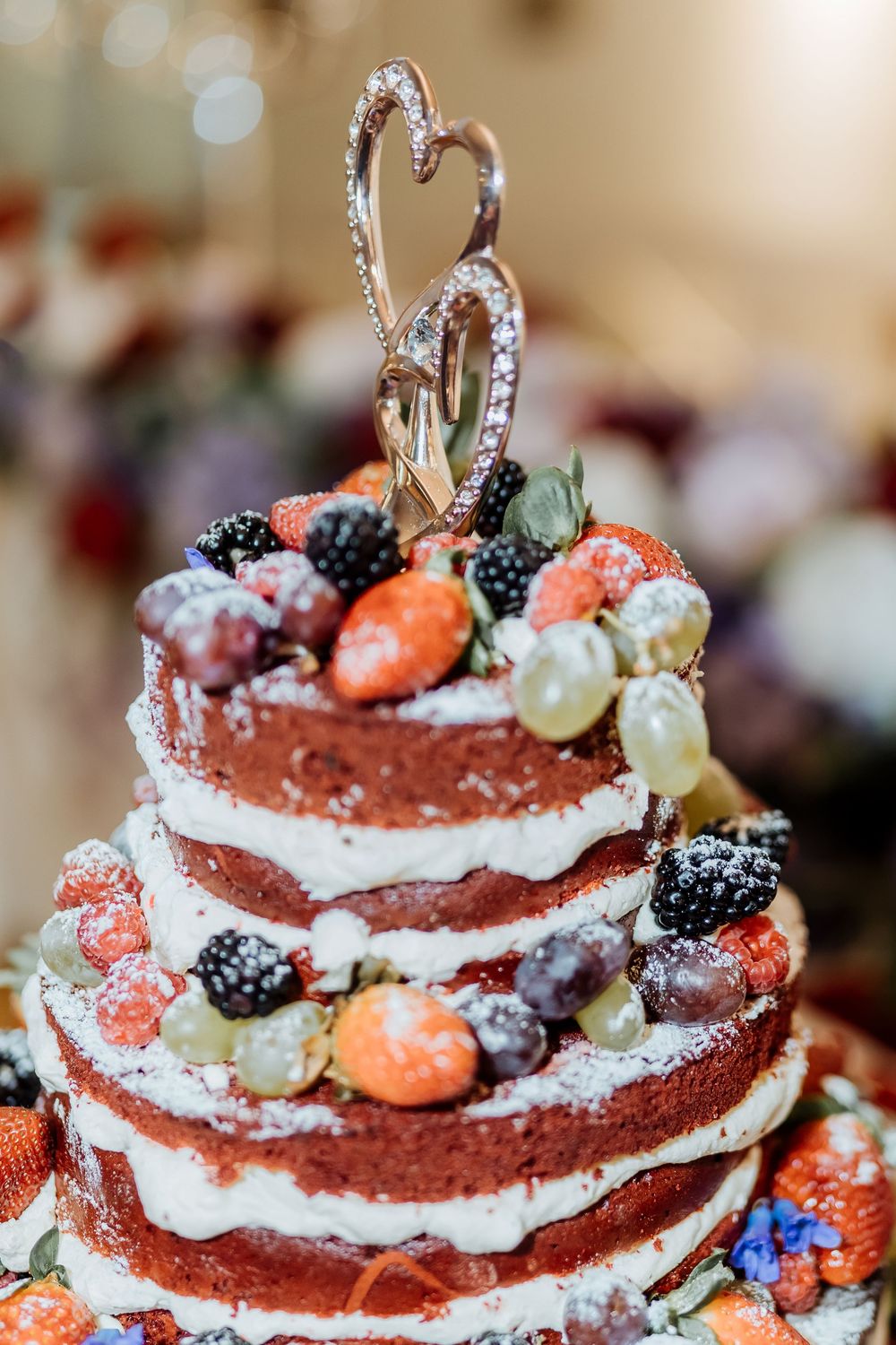 Rustic naked wedding cake decorated with fresh berries and topped with a silver heart ornament.