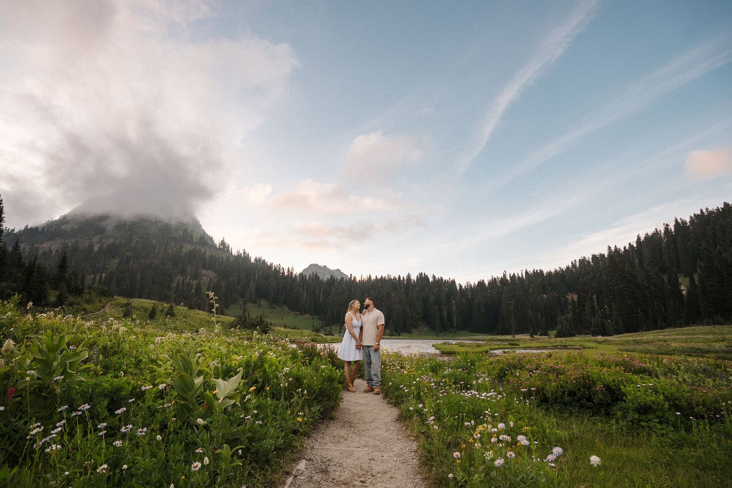 A scenic hiking trail through a wildflower meadow leads toward misty mountain peaks under blue summer skies.