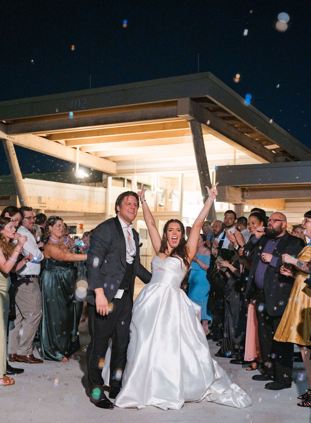 Wedding couple exits venue during nighttime celebration while guests blow bubbles and cheer under illuminated overhang.