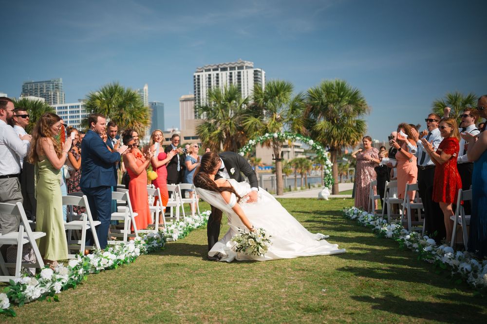 A couple shares their first kiss at a romantic outdoor wedding ceremony with palm trees and guests in coral attire.