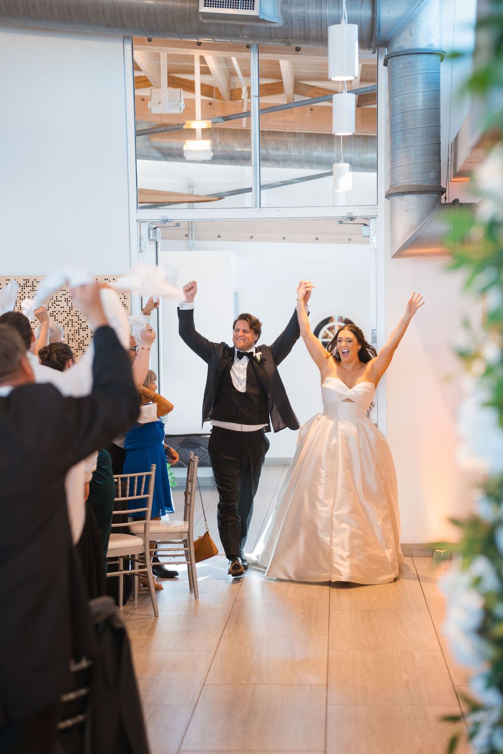Wedding couple and guests celebrate with joyful dancing in a bright, modern reception venue with white decor.