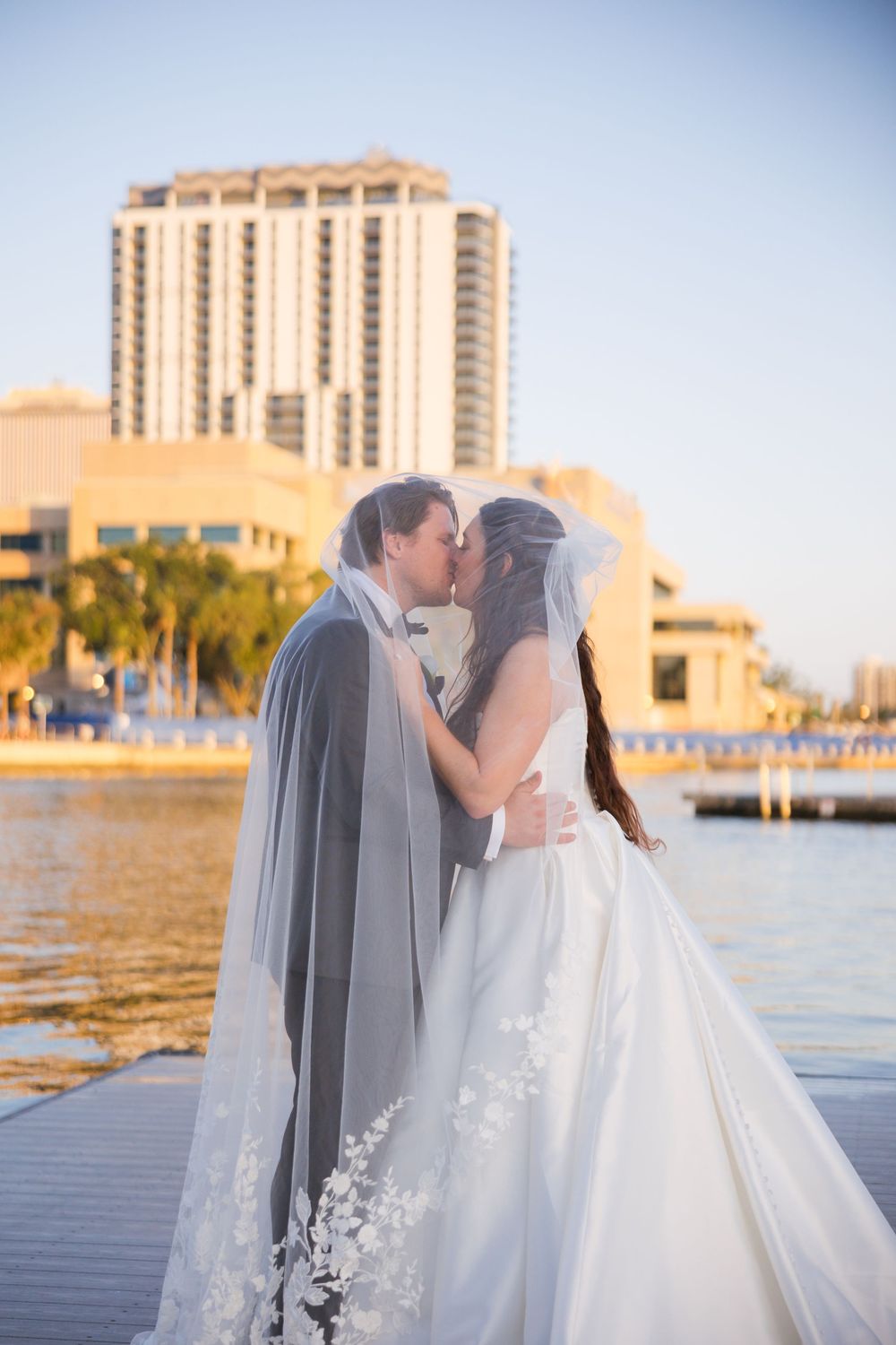 A wedding couple shares a romantic moment at sunset on a waterfront dock with a high-rise building in the background.
