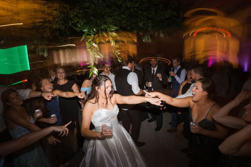 A bride in a white wedding dress dances on a crowded dance floor at a reception with colorful lighting effects.