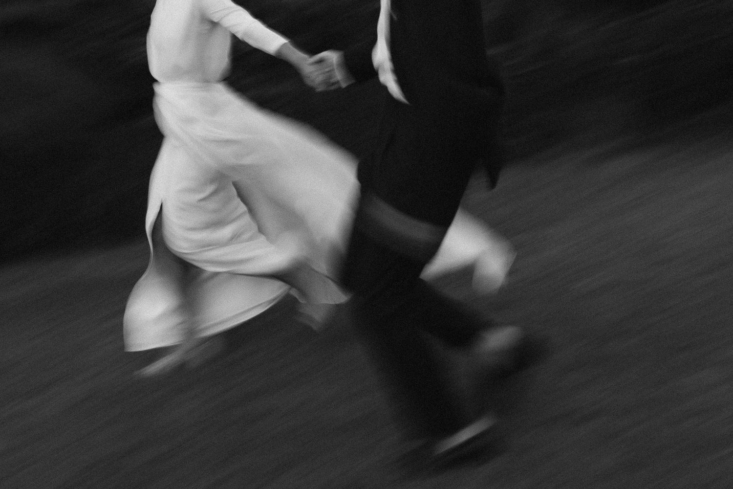 Black and white motion blur photograph showing legs and feet of dancers in motion on a dark floor.