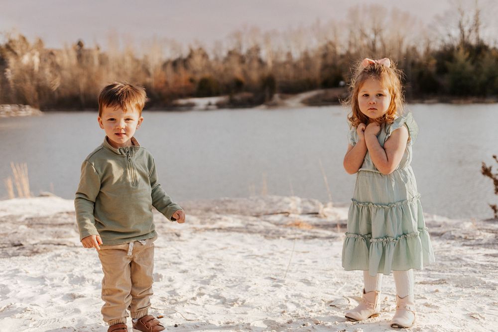 Two young children pose outdoors near a frozen lake, boy in green sweater and girl in mint dress on snowy ground.