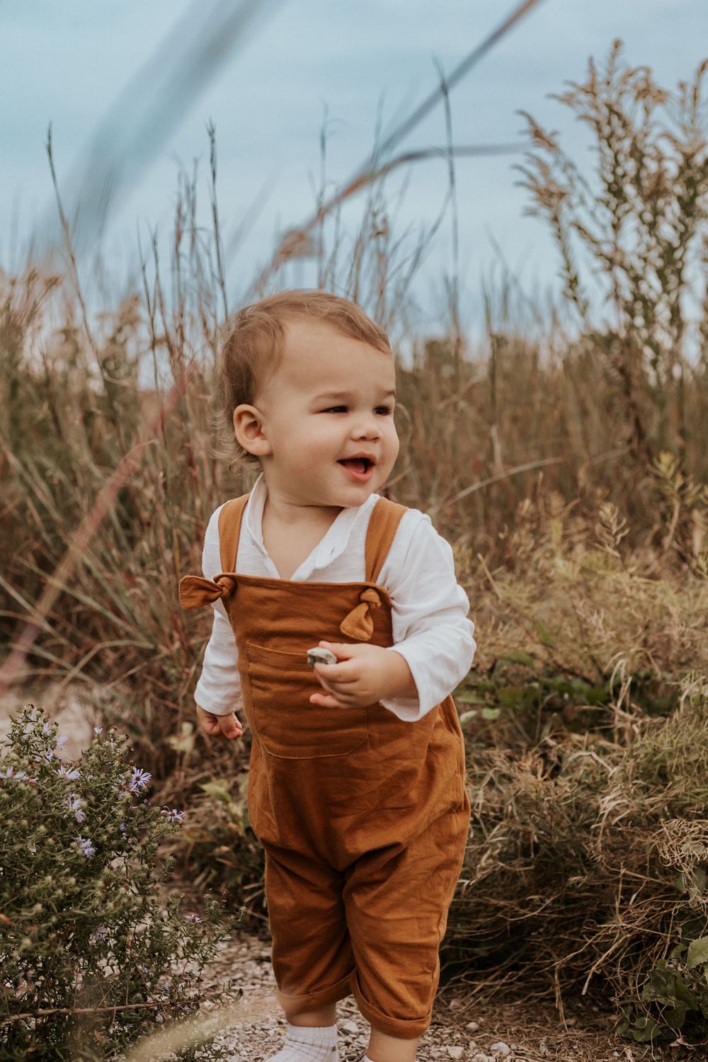 Smiling toddler in brown overalls and white shirt standing in a field of wildflowers and tall grass.