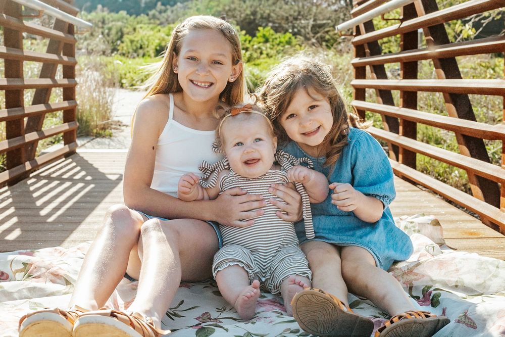 Three happy children sitting together on a blanket on a wooden bridge outdoors on a sunny day.