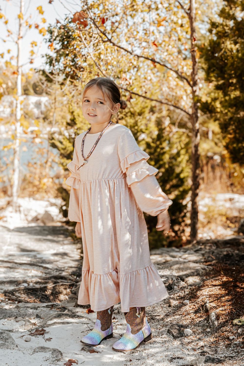 A young girl in a blush pink ruffle dress and holographic shoes poses outdoors among autumn trees.