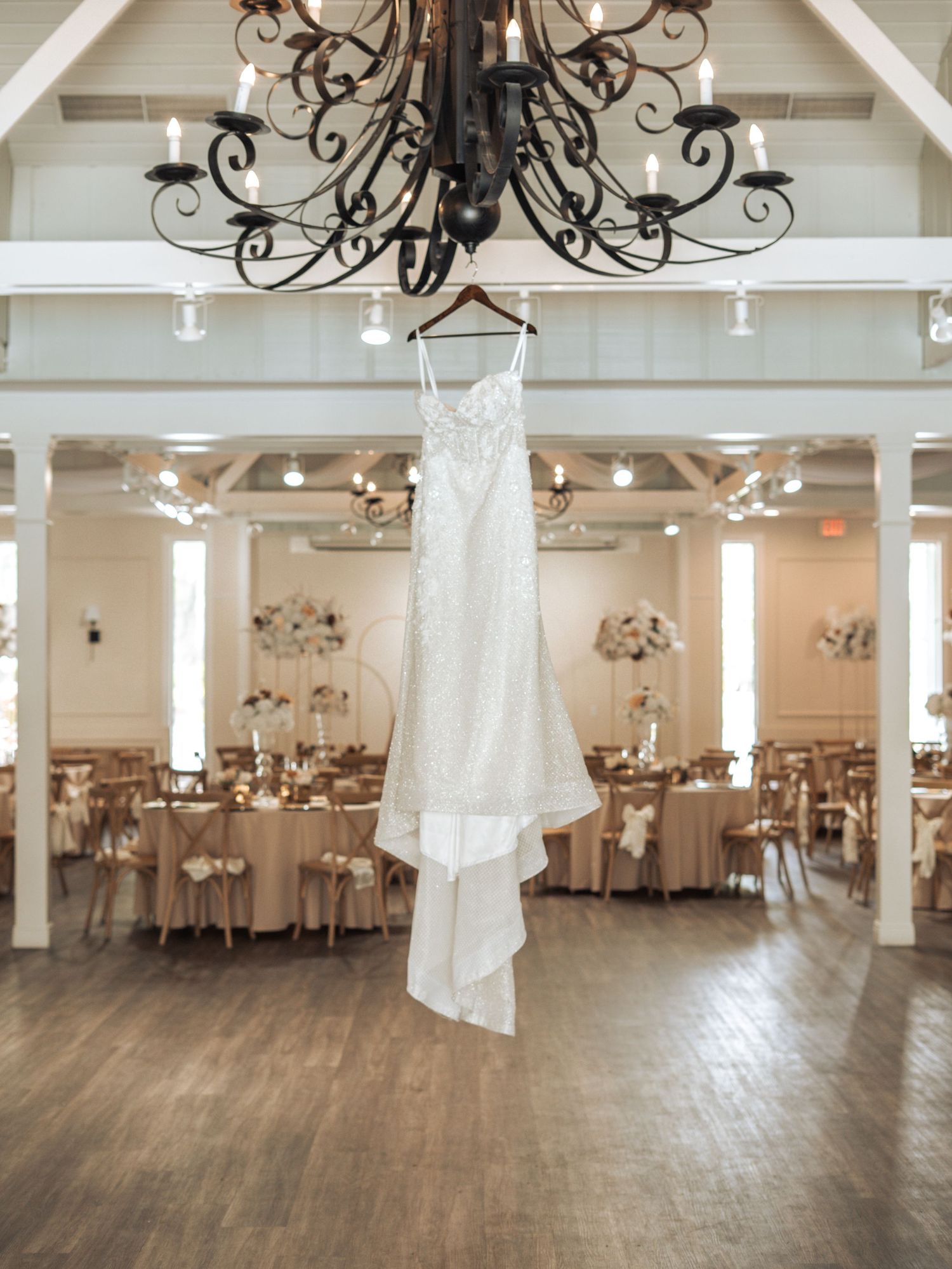 A white wedding dress hangs from an ornate chandelier in an elegant reception venue with round tables and hardwood floors.