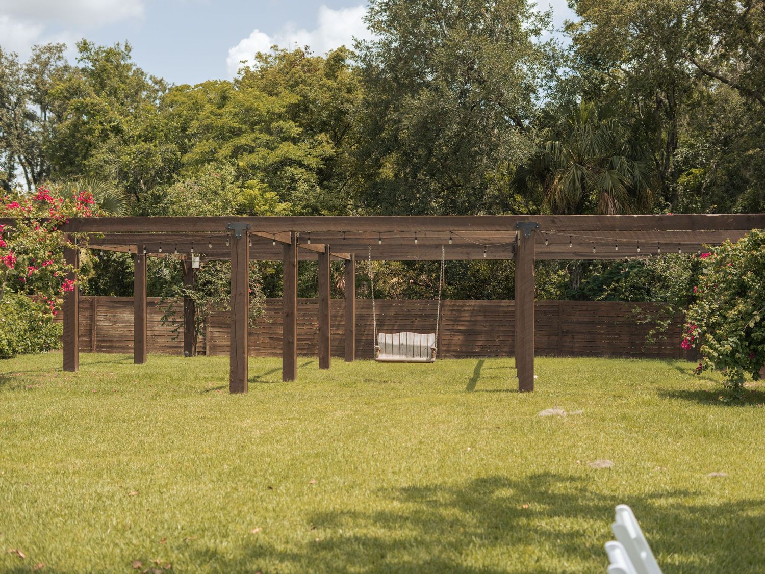 A wooden pergola with a white porch swing stands in a sunny garden surrounded by lush green trees and grass.