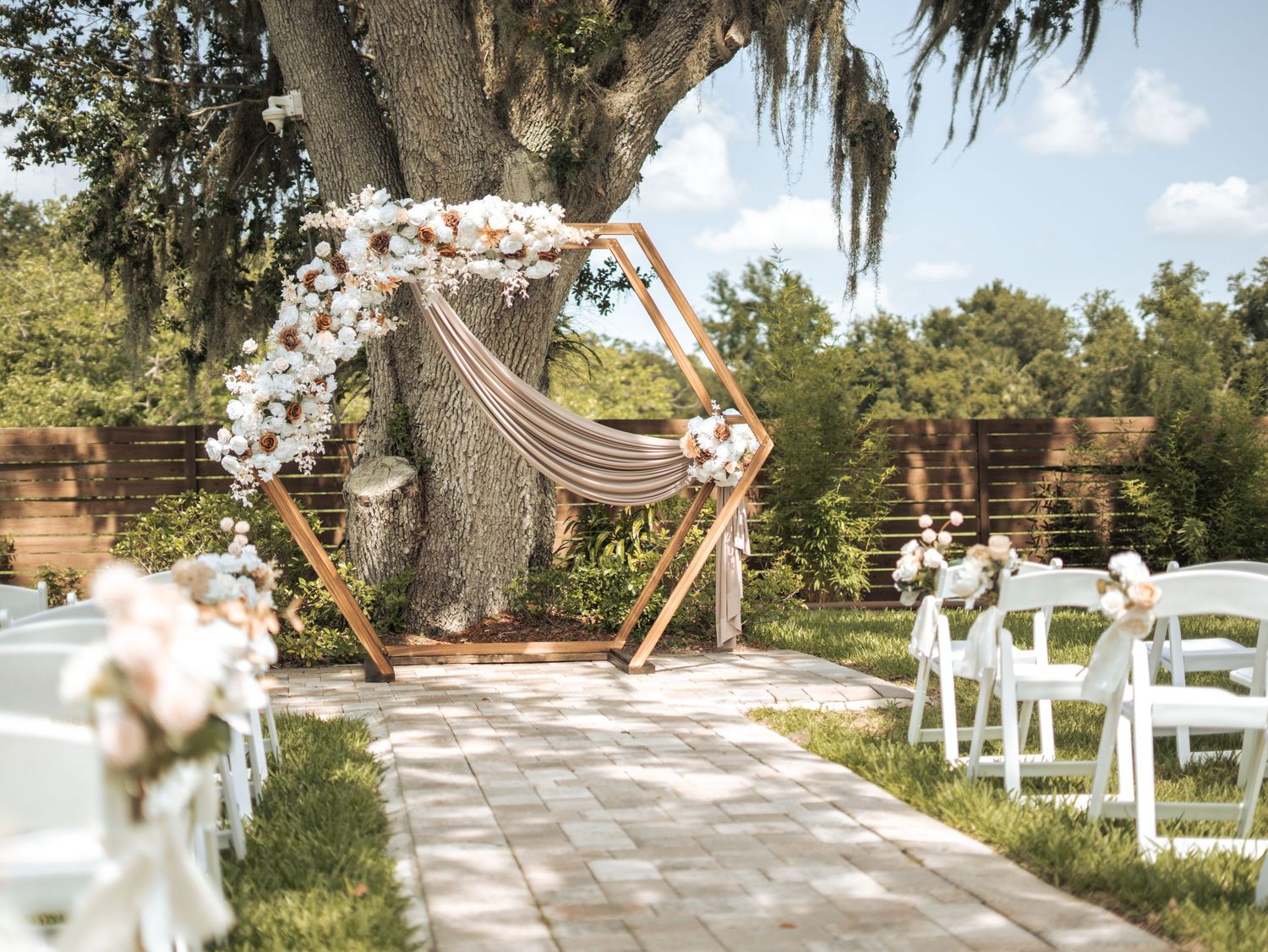 A rustic outdoor wedding ceremony setup with white chairs and floral arrangements under Spanish moss-draped trees.