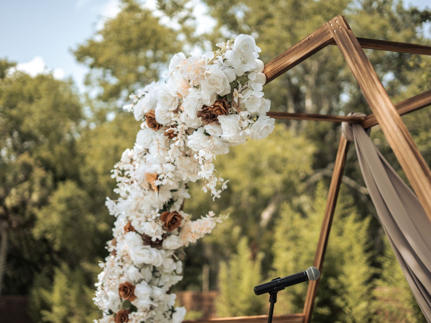 White floral garland decorating a geometric copper or rose gold arch frame in an outdoor garden setting.