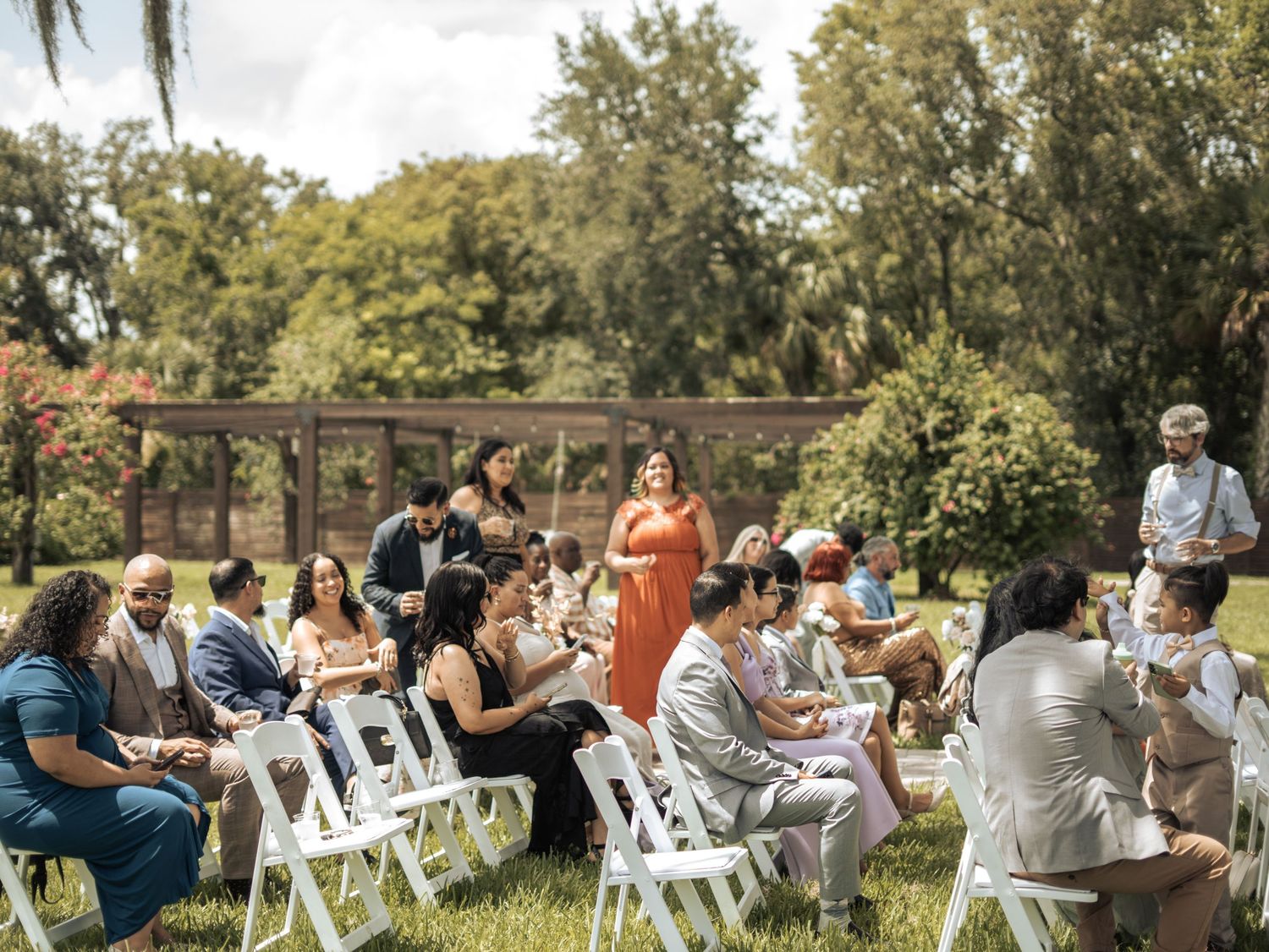 Guests attend an outdoor garden wedding ceremony with white chairs setup on grass under a wooden pergola structure.