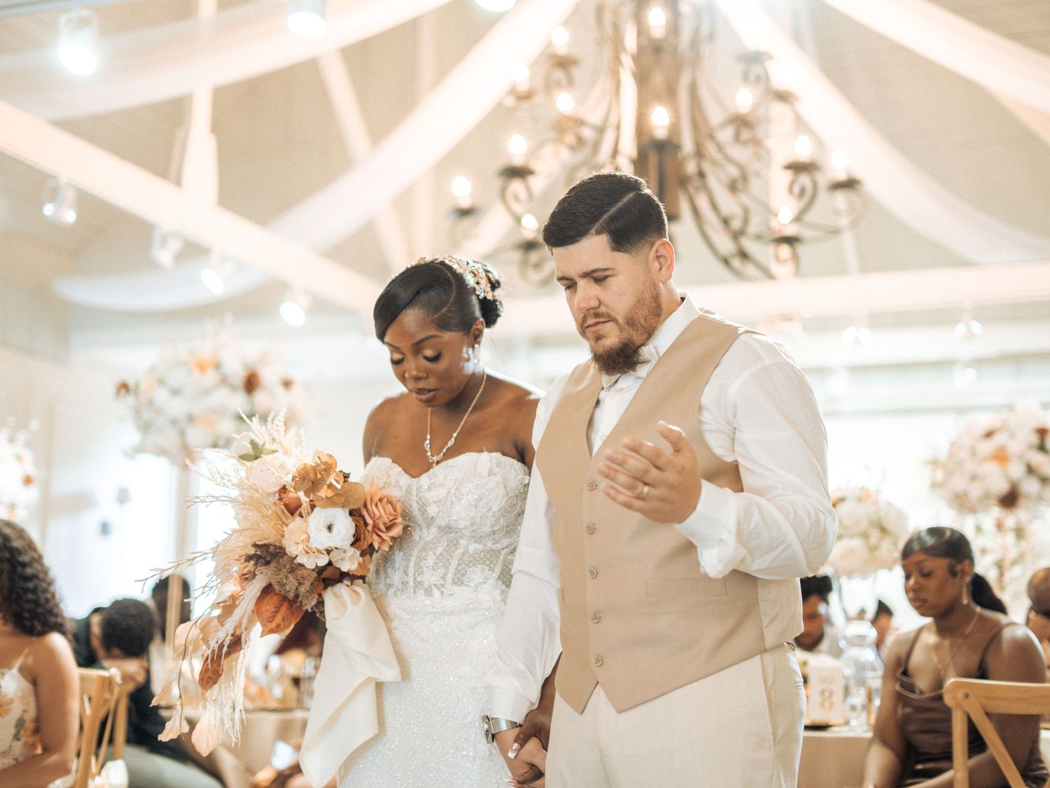 A couple prays together during their beautiful wedding reception in an elegant white-draped room with chandeliers.