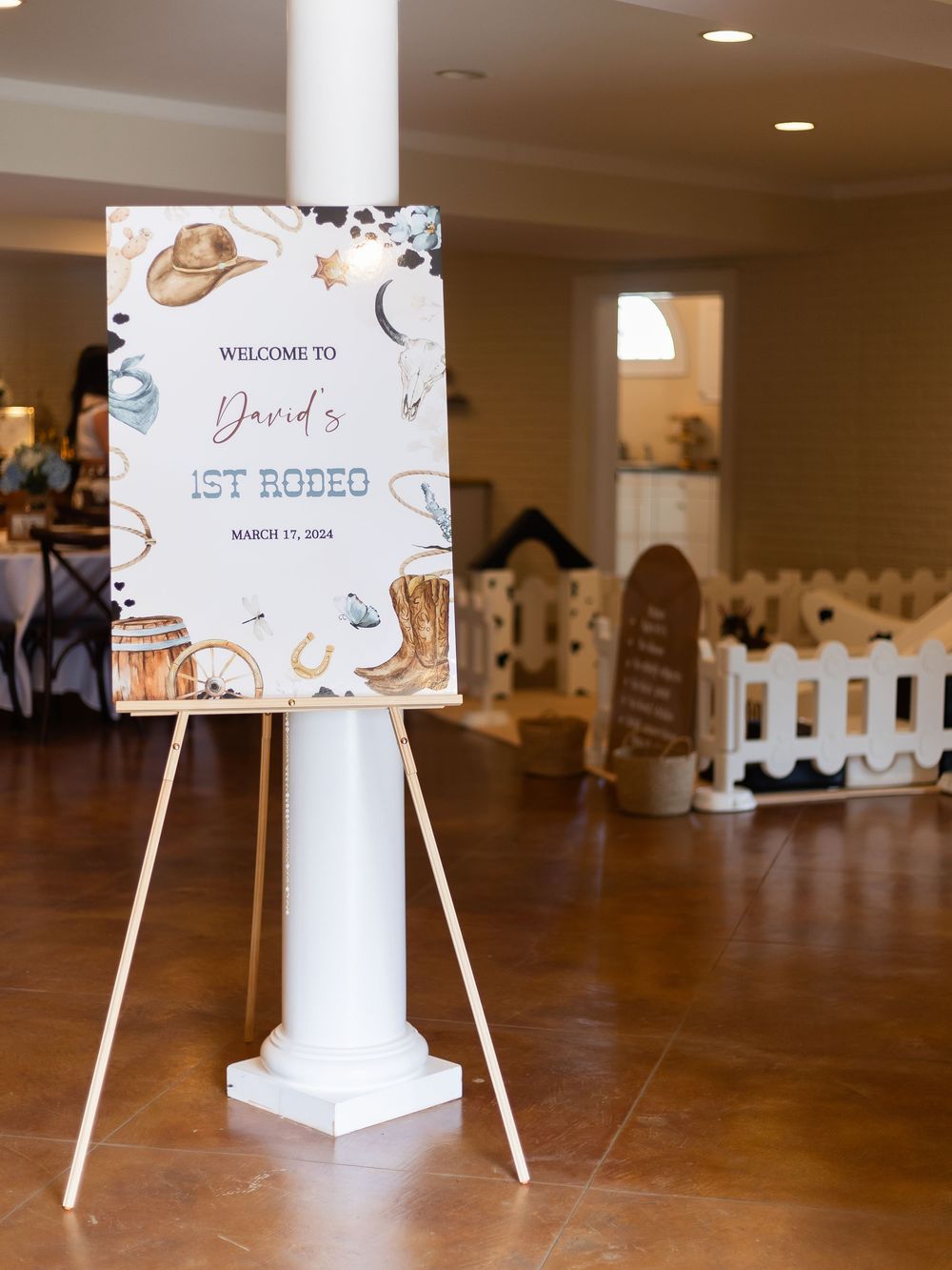Welcome sign for first rodeo event displayed on white easel with brown decor and white picket fence in background.
