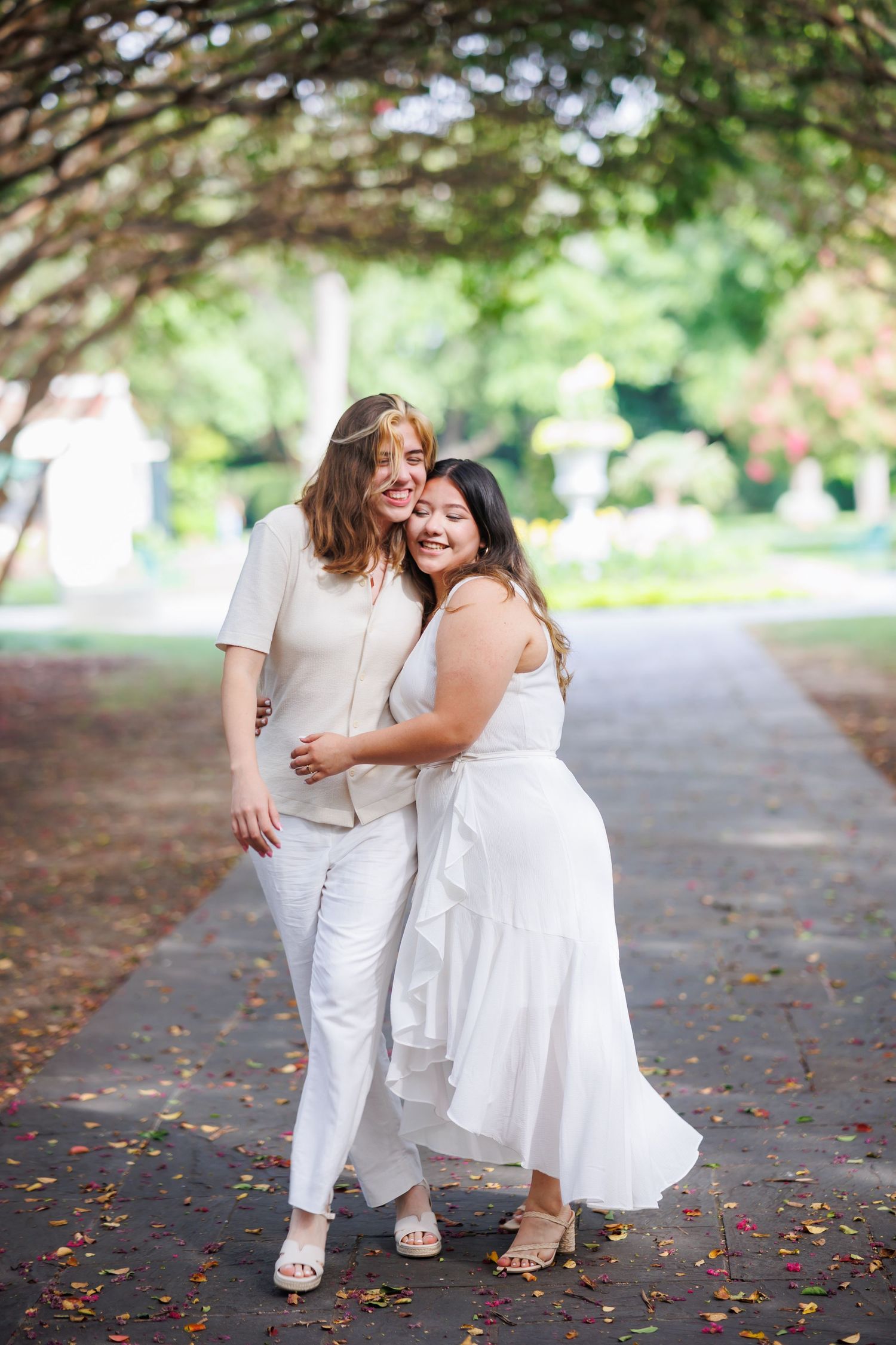 Two women in white dresses hold hands while walking down a tree-lined path during their engagement photoshoot at Dallas Arboretum
