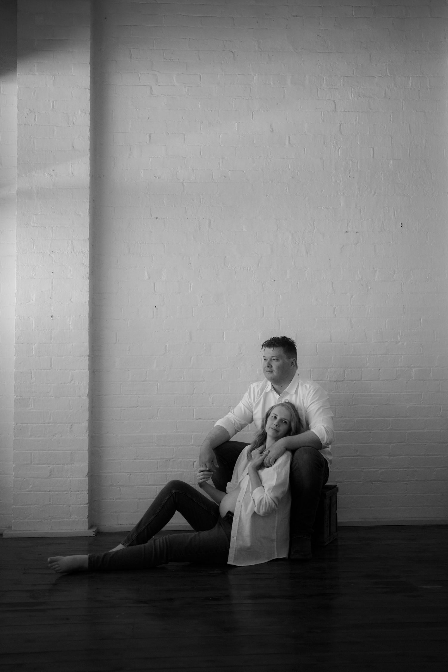 Black and white photo of a couple in white shirts, woman sitting on floor leaning on seated man against brick wall.