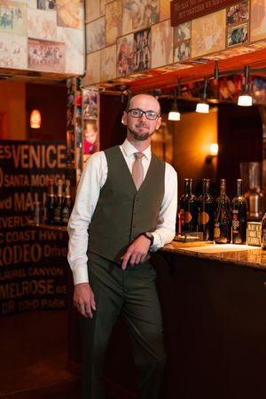 Professional bartender in formal attire at an upscale bar with vintage wall decor and bottles displayed.