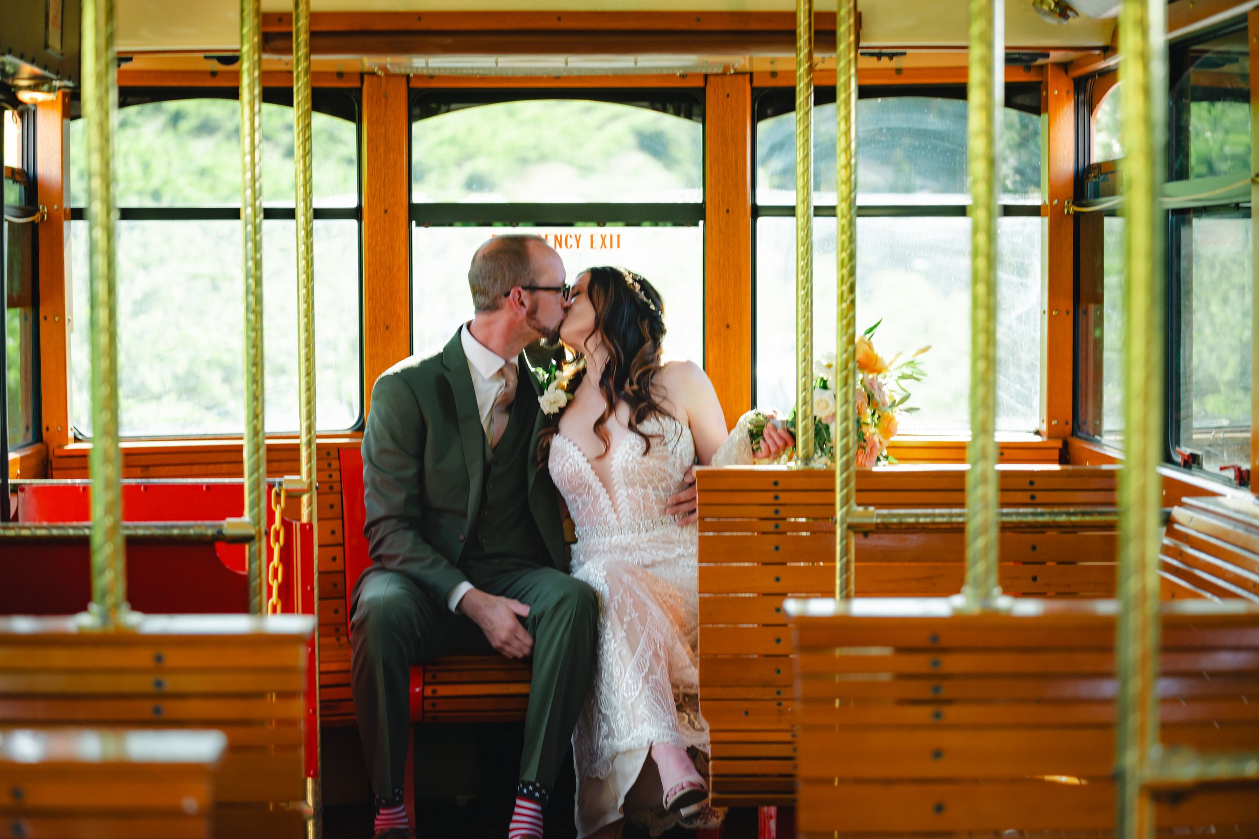 Wedding couple embraces on classic vintage trolley car while peering through windows during romantic moment.
