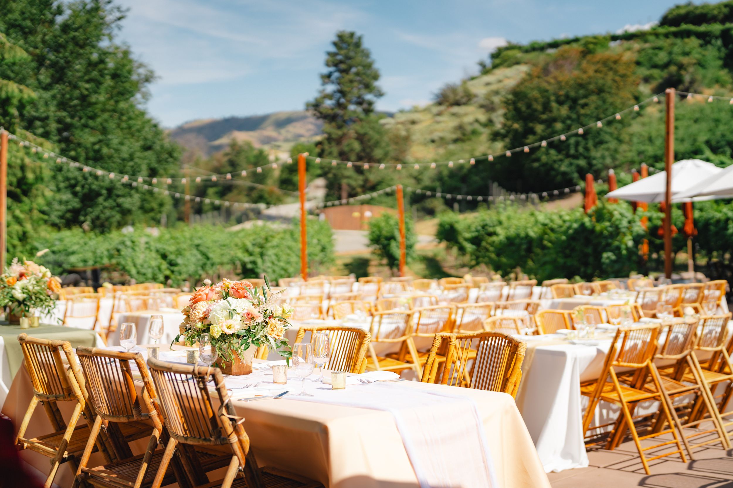 Wedding reception tables with white linens and wooden folding chairs set up outdoors with mountain scenery in the background.