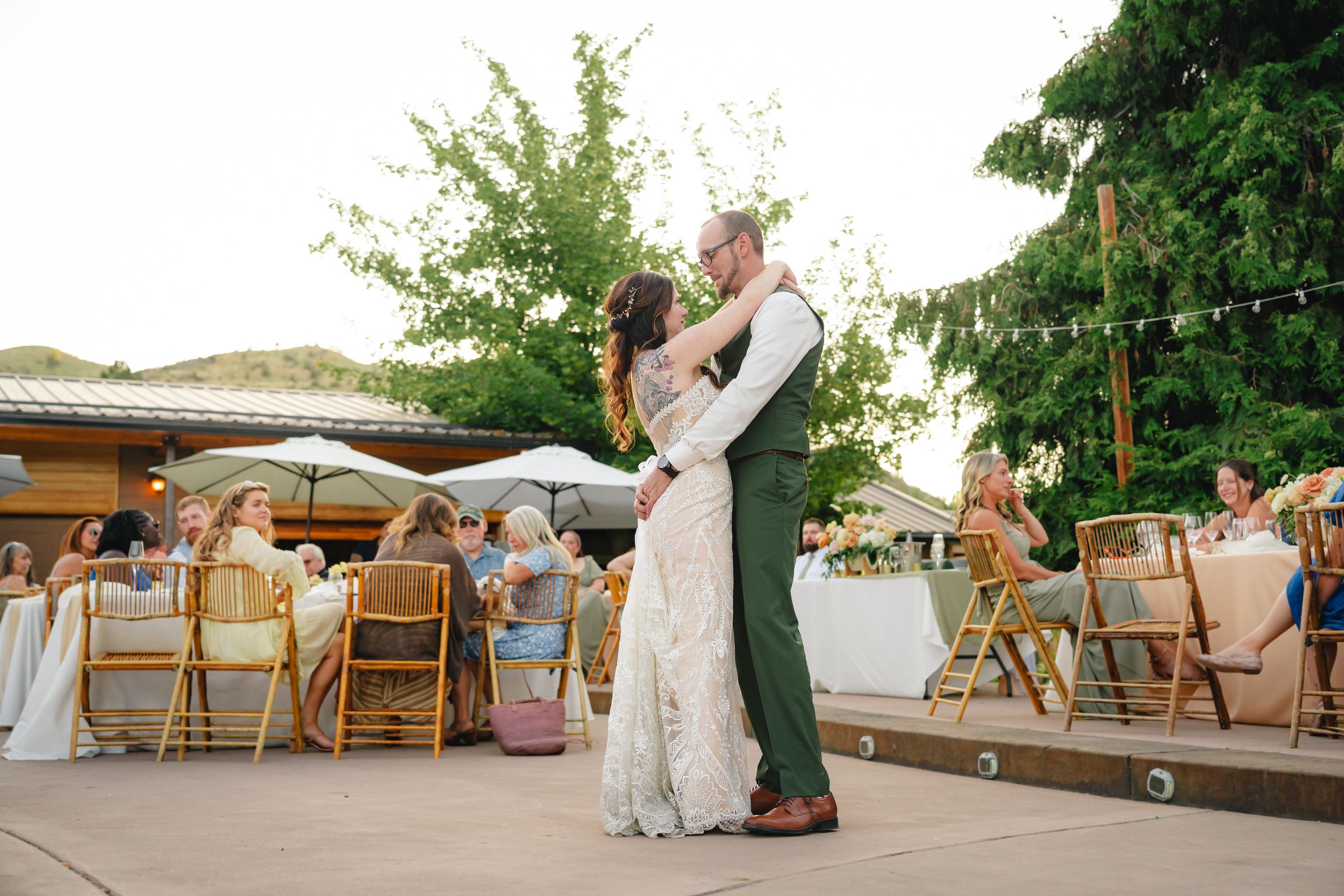A couple shares their first dance at an outdoor wedding reception with wooden folding chairs and greenery in the background.