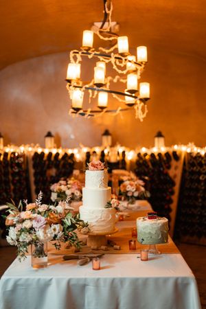 Wedding cake display table with multiple tiers and floral decorations illuminated by warm candlelight and chandeliers.