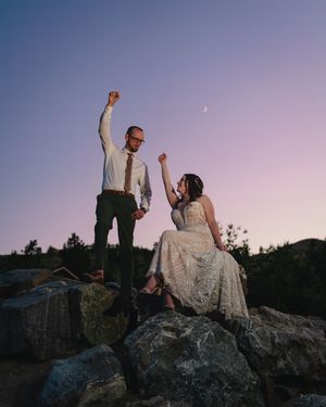 Newlyweds celebrate joyfully on rocky cliffs at sunset with a purple sky backdrop.