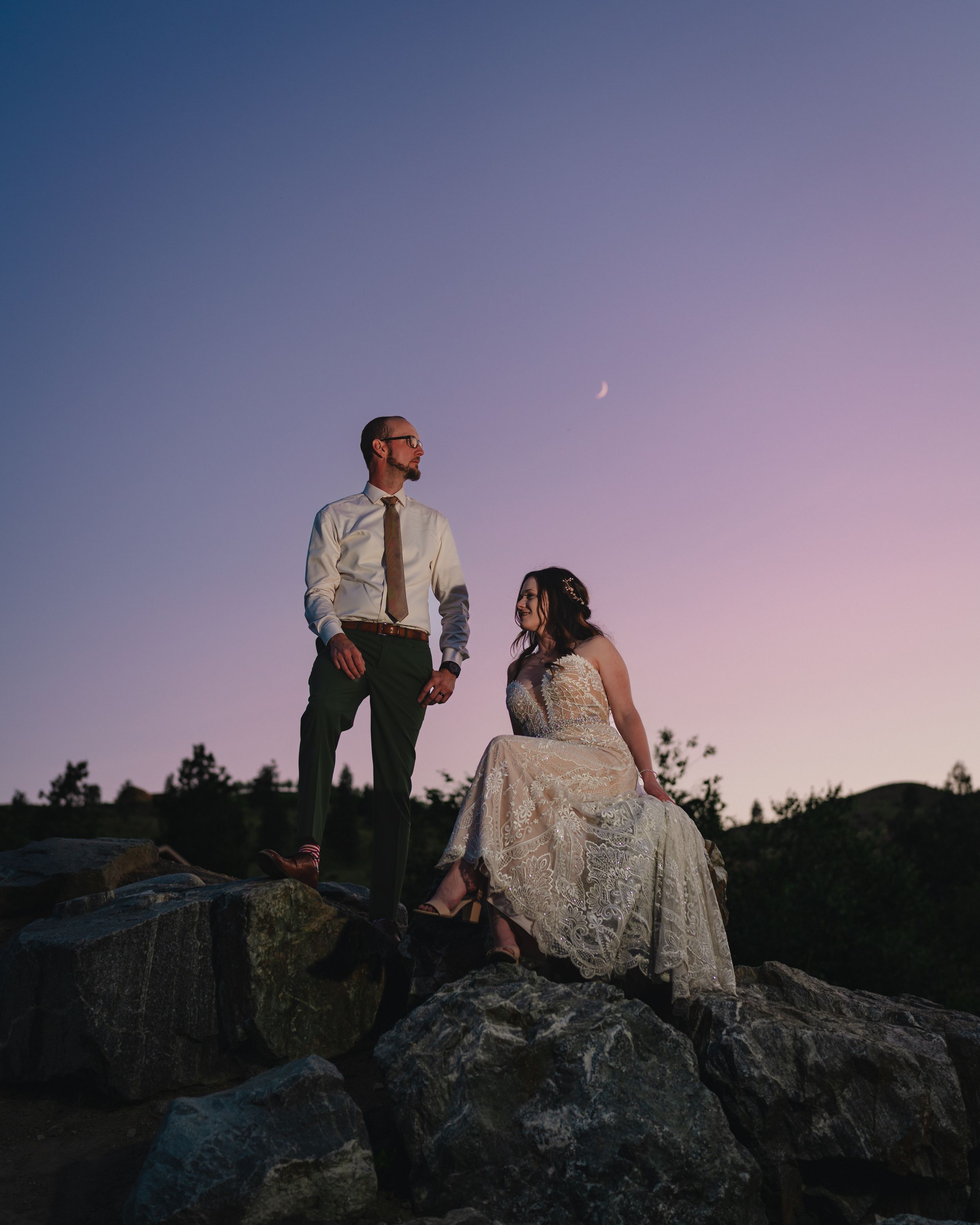 Wedding couple sits on coastal rocks against a dusky purple evening sky.