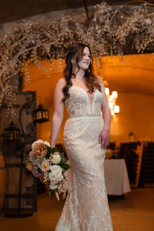 A bride in a detailed lace wedding gown holds a blush and cream bouquet while standing in an elegantly lit venue.