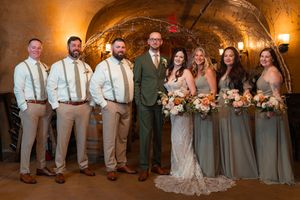 Wedding party poses in rustic wine cellar setting with bridesmaids in sage dresses and groomsmen in tan suspenders.