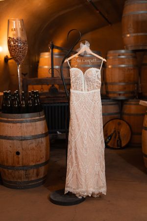 Elegant white lace wedding dress displayed in rustic wine cellar setting with wooden barrels and warm lighting.