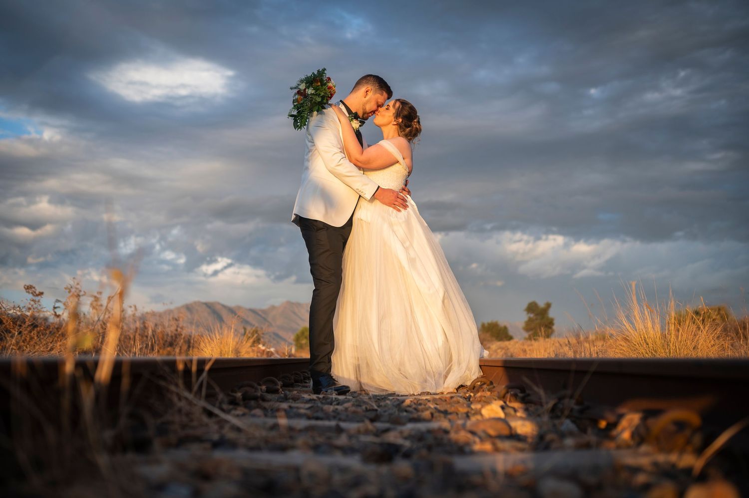 A romantic sunset wedding photo with dramatic cloudy sky and desert mountains in the background.