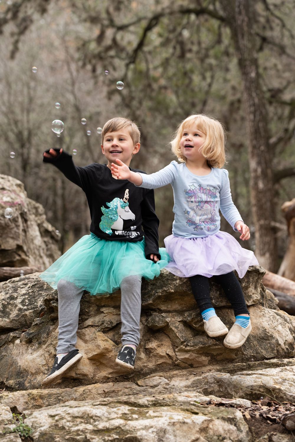 Two young children in tutus sitting on rocks and playing with bubbles in a wooded outdoor setting.