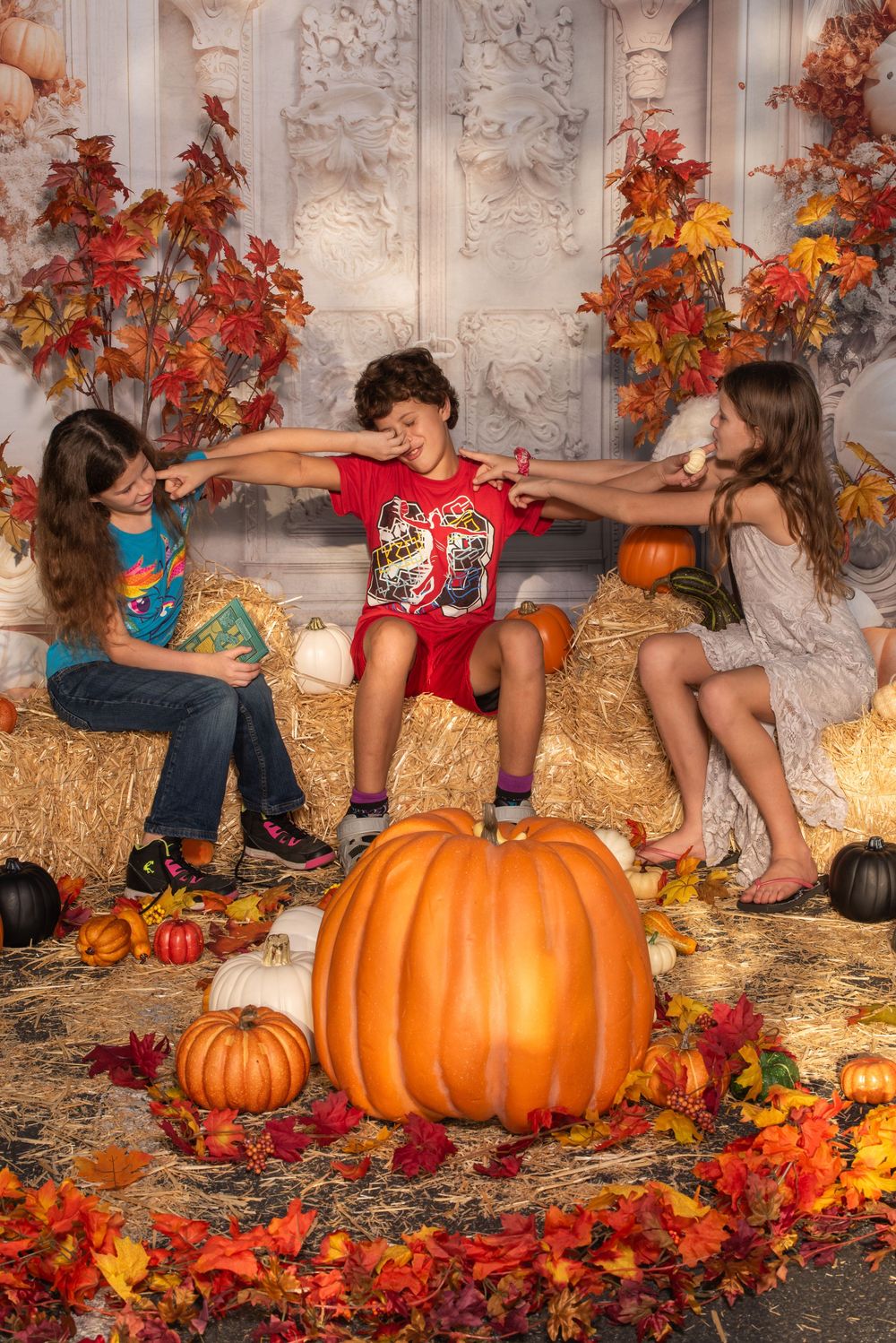 Children sitting around large orange pumpkins with autumn leaves and hay bales in a festive fall photo setting.