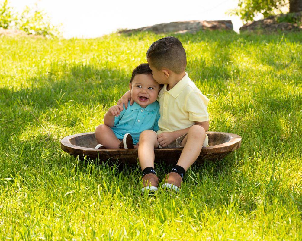 Two children share a playful moment while sitting together in a saucer sled on sunny grass.