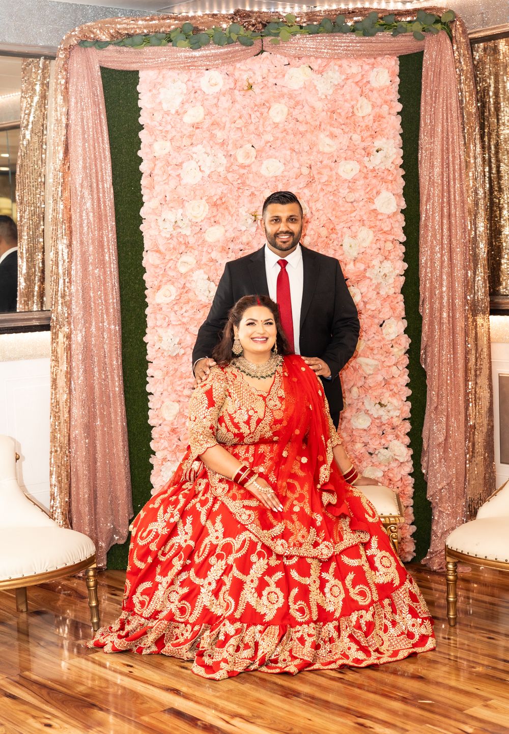 A couple poses in front of a pink floral backdrop, with the woman wearing a traditional red and gold lehenga.