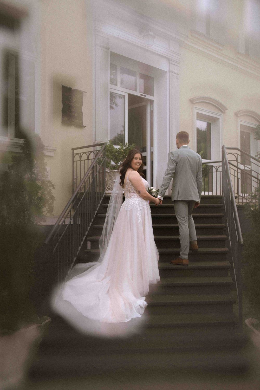 Bride in flowing white gown stands with groom on formal building steps.