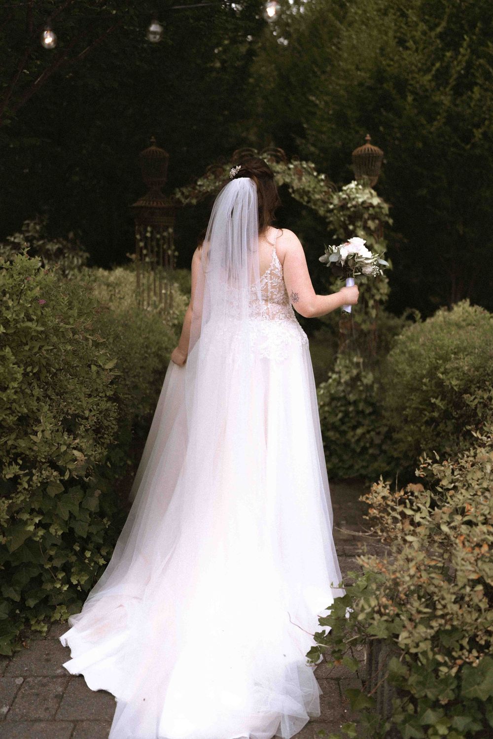 Back view of wedding gown with flowing train and veil in garden setting with string lights.