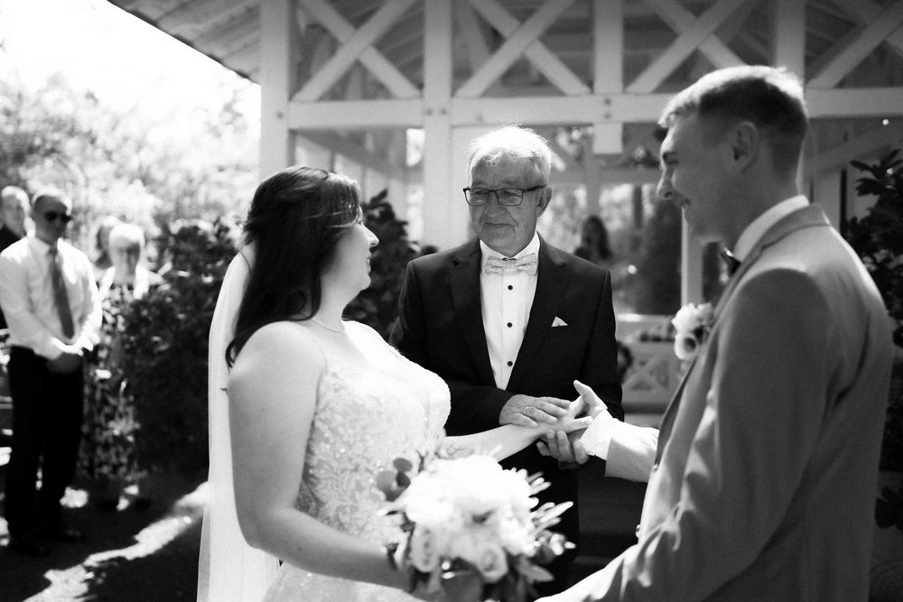 An outdoor wedding ceremony takes place under a white wooden pergola structure.