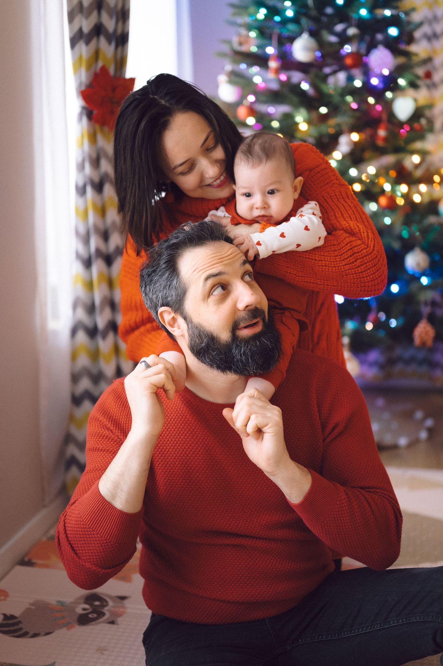 Parent in red sweater playing peek-a-boo in sequence with baby by Christmas tree with twinkling lights.