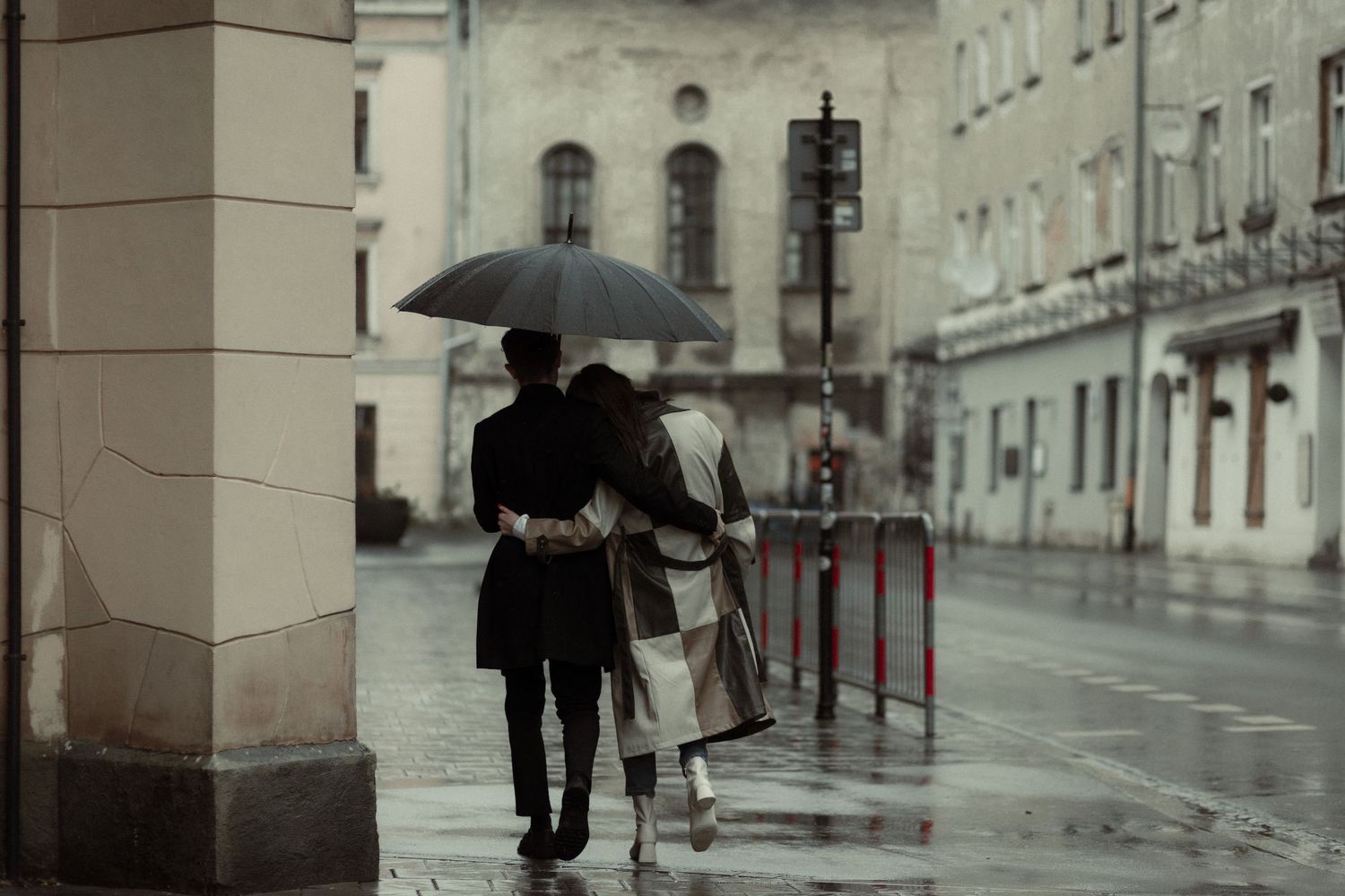 People walk with umbrellas down a rain-soaked cobblestone street in a historic European city during gloomy weather.