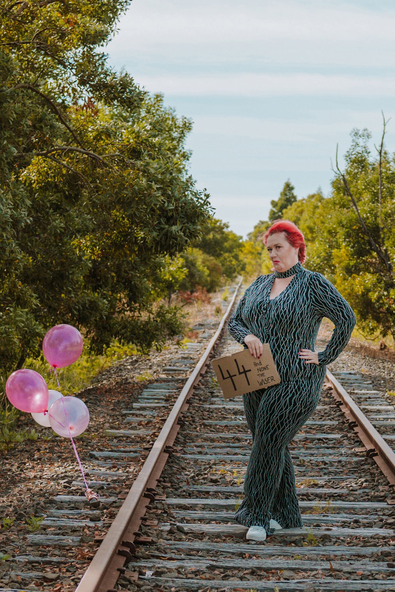A person in a striped dress stands on railroad tracks holding pink balloons against a leafy green background.