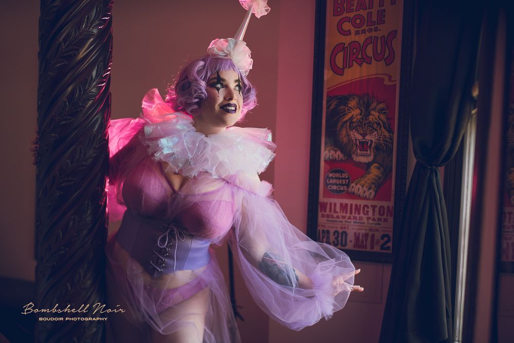 A burlesque performer in a pink and white sheer costume poses dramatically against a darkened backstage area with vintage posters.