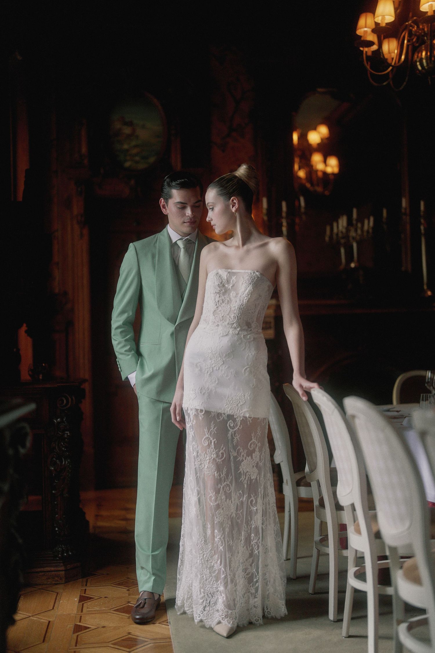 A couple in wedding attire pose together in a dimly lit vintage room with chandeliers and white chairs.