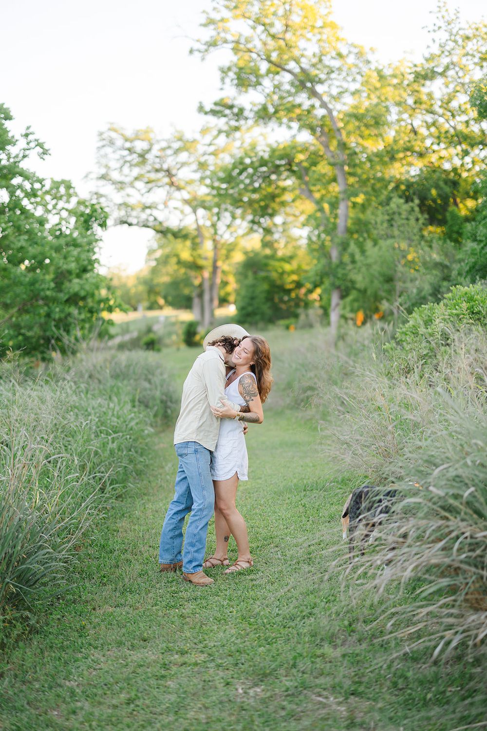 A couple shares a romantic moment on a path surrounded by tall grass and autumn trees in warm sunlight.