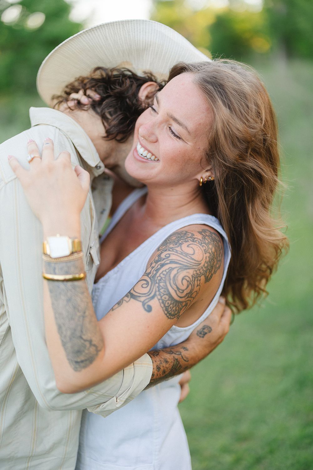 A laughing person with tattoos embraces another in a candid outdoor moment, wearing a blue tank top.