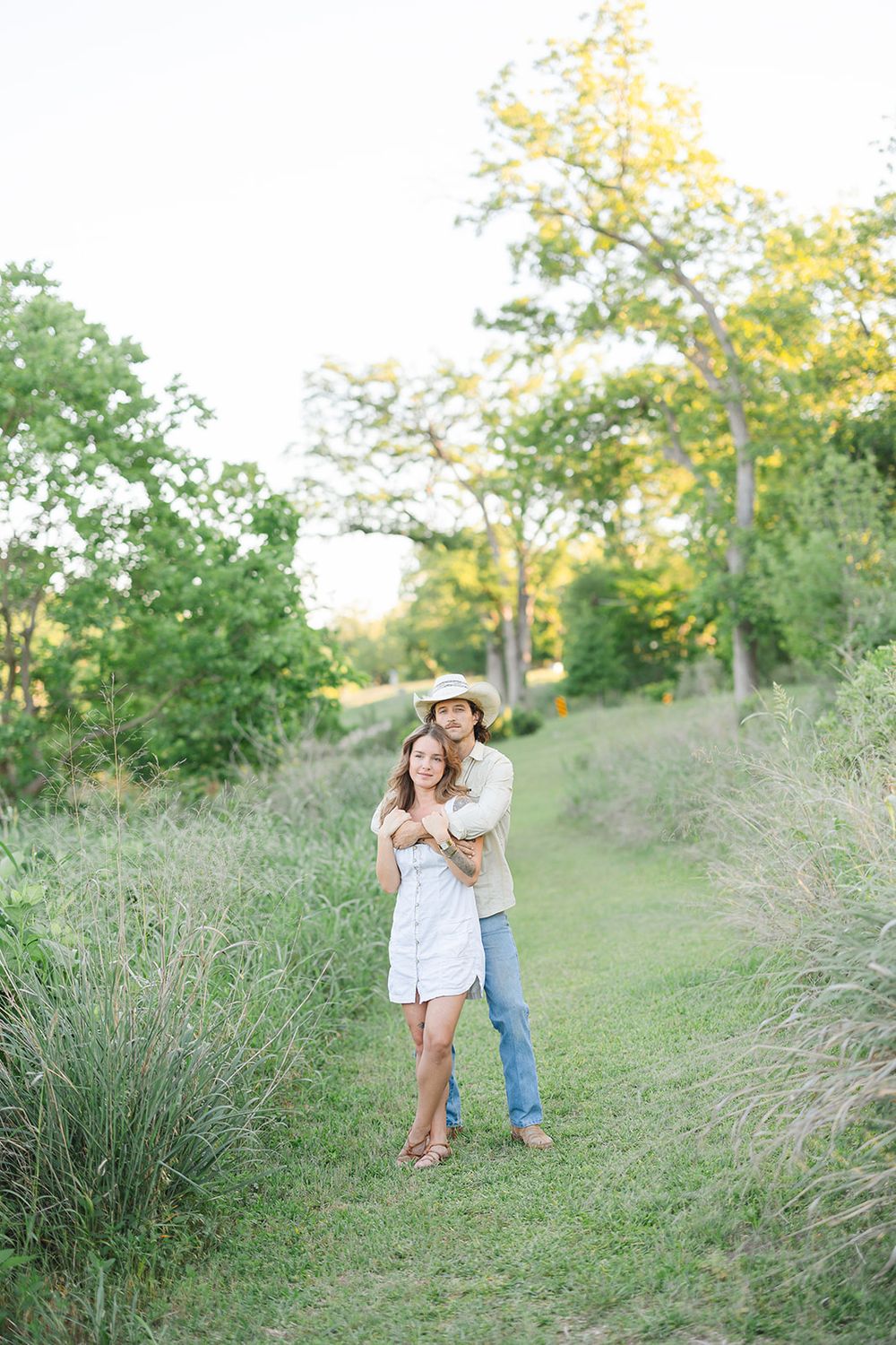 Romantic couple playfully embraces in tall grass field during golden hour among trees at sunset.