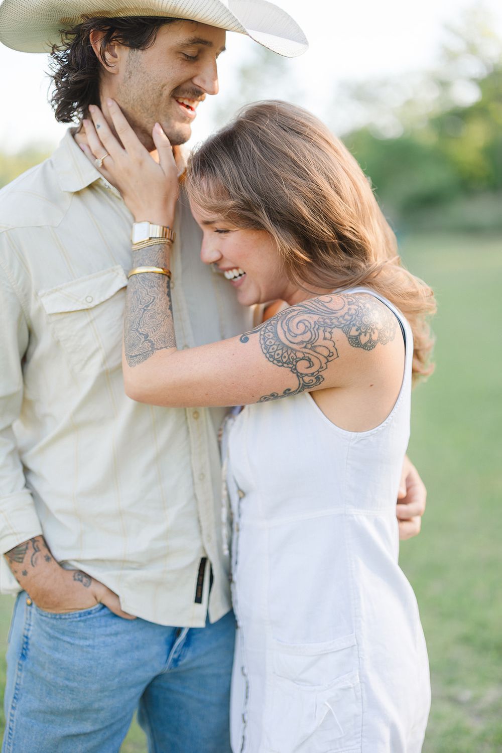 Romantic couple embracing outdoors with woman in white dress showing intricate arm tattoo and man wearing casual attire.